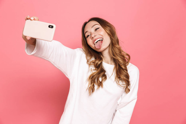 Portrait of a happy young woman taking selfie with mobile phone isolated over pink background, sticking her tongue out - 写真・画像