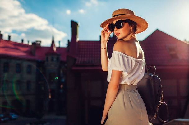 Giovane donna elegante indossa cappello e occhiali da sole all'aperto. Elegante ragazza con zaino e telefono a piedi a Leopoli indossando abito retrò
 - Foto, immagini