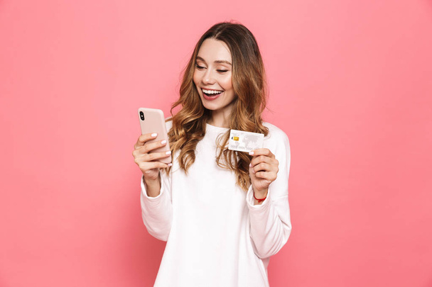 Portrait of a happy young woman using mobile phone, holding plastic credit card isolated over pink background - Foto, immagini