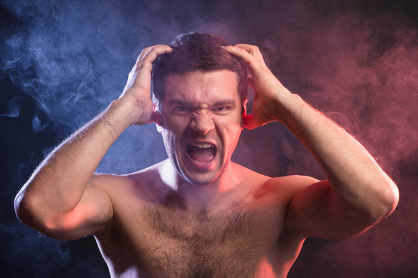 Man grabs his head with his hands and screams from overexertion. Negative emotions of a person. Studio portrait on a black background of a young man of Caucasian appearance. The concept of hysteria and stress - Photo, Image