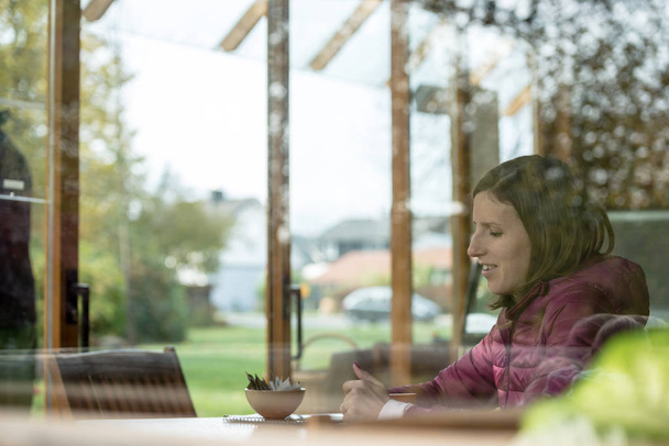 Vue à travers une fenêtre vitrée d'une jeune femme assise dans un café seule
. - Photo, image