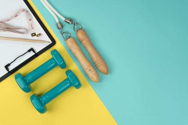 top view of the green dumbbells and jump rope, vinyl tape measure and black clipboard with pencil on the yellow and blue background - Fotoğraf, Görsel