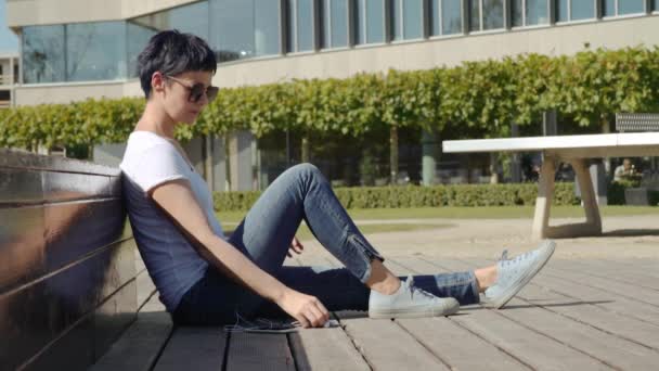 business woman in a white T-shirt sits in front of an office building and listens to music - Záběry, video