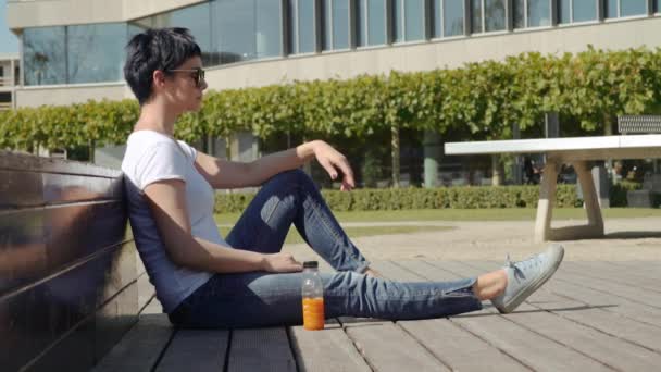 woman in a white T-shirt sitting in front of an office building and drinking orange juice - Materiaali, video