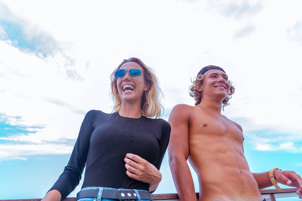 Χαμογελώντας νεαρό ζευγάρι ενεργό surfers έχοντας διασκέδαση στην παραλία μετά τον αθλητισμό. Όμορφη Θαλασσογραφία στο φόντο - Φωτογραφία, εικόνα