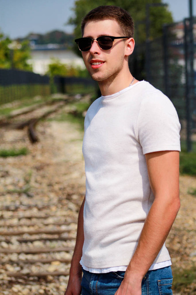 Handsome young guy with sunglasses posing and enjoying a nice sunny day. Urban city environment. Old railway in background. Male model photo shoot, urban fashion, lifestyle. White T-shirt and jeans. - Foto, Bild