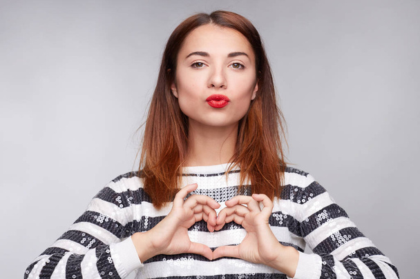 Body language concept. Stylish beautiful female makes heart gesture, looks seriously at camera, wears glitter blouse and red lipstick, expresses peace and love , isolated over gray studio background.  - Photo, Image