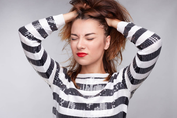 Stressful young cute female has terrible headache, keeps both hands on head, clenches teeth, wears black and white long sleeved shirt, has makeup, red lips, suffers against gray studio background. - Photo, Image
