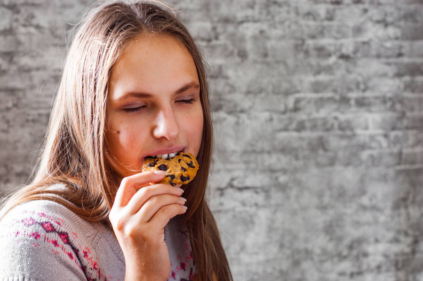 portrait of young teenager brunette girl with long hair eating cookie or biscuit on gray wall background - Fotó, kép