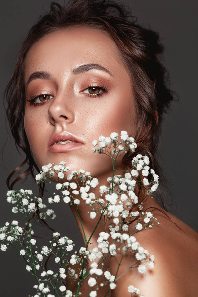 Glamour portrait of beautiful woman model with fresh daily makeup and romantic wavy hairstyle.  Fashion shiny highlighter on skin, sexy gloss lips make-up and dark eyebrows. Cute freckles on young face. Sensual woman with white flowers. - Foto, immagini