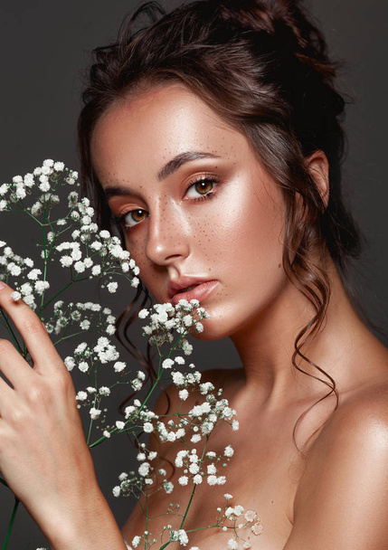 Glamour portrait of beautiful woman model with fresh daily makeup and romantic wavy hairstyle.  Fashion shiny highlighter on skin, sexy gloss lips make-up and dark eyebrows. Cute freckles on young face. Sensual woman with white flowers. - Photo, image