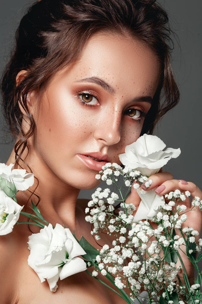 Glamour portrait of beautiful woman model with fresh daily makeup and romantic wavy hairstyle.  Fashion shiny highlighter on skin, sexy gloss lips make-up and dark eyebrows. Cute freckles on young face. Sensual woman with white flowers. - Foto, Imagen