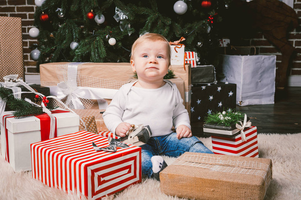 Kompozycja zima i święta Bożego Narodzenia. Dziecko chłopiec kaukaski blond 1 roku życia, siedzi na piętro w pobliżu choinkę z wystrój nowy rok na dywan shaggy skóry odbiera prezenty, otwiera pudełeczka wieczorem. - Zdjęcie, obraz