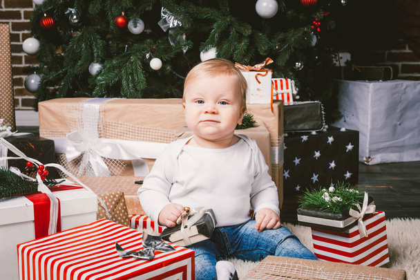 θέμα χειμώνα και τις διακοπές των Χριστουγέννων. Παιδί αγόρι Ξανθιά καυκάσιος 1 χρονών κάθονται σπίτι πάτωμα κοντά στο χριστουγεννιάτικο δέντρο με το νέο έτος διακόσμηση στο δέρμα δασύτριχος χαλί λαμβάνει δώρα, ανοίγει κουτιά δώρων βράδυ. - Φωτογραφία, εικόνα