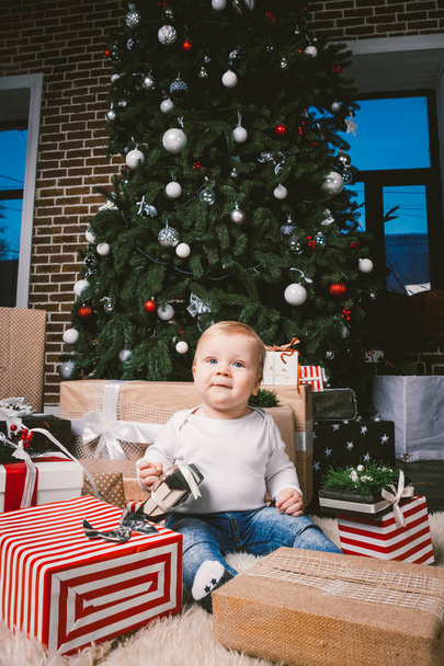 Kompozycja zima i święta Bożego Narodzenia. Dziecko chłopiec kaukaski blond 1 roku życia, siedzi na piętro w pobliżu choinkę z wystrój nowy rok na dywan shaggy skóry odbiera prezenty, otwiera pudełeczka wieczorem. - Zdjęcie, obraz