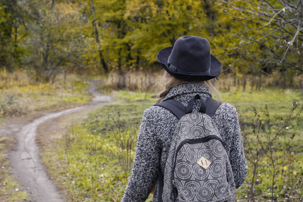 Girl with a backpack and a hat standing in a autumn forest. Back view - Фото, зображення
