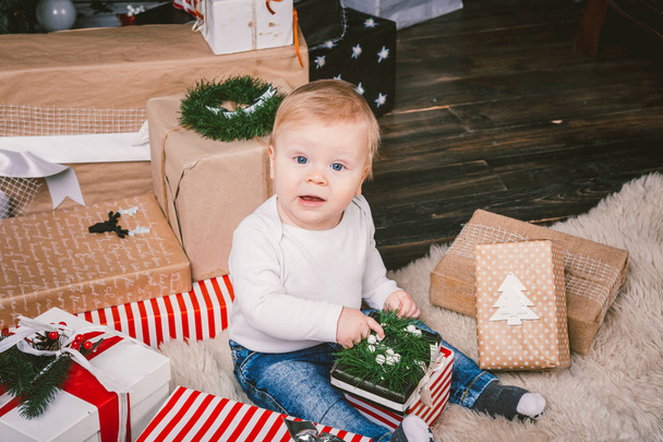 theme winter and Christmas holidays. Child boy Caucasian blond 1 year old sitting home floor near Christmas tree with New Year decor on shaggy carpet skin receives gifts, opens gift boxes in evening. - Photo, Image