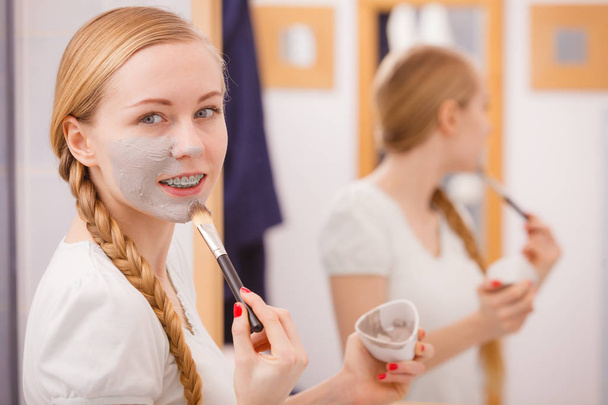Facial dry skin and body care, complexion treatment at home concept. Happy young woman applying grey mud mask on her face with brush. - 写真・画像