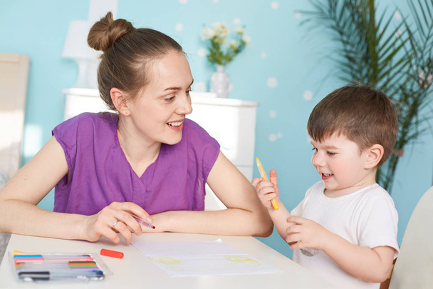 Cheerful mum and son paint together, sit at table, like creativity, spend free time at home, use colourful pencils, look positively at each other. Education, children, family and handcraft concept - Foto, afbeelding
