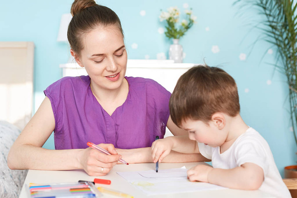 Attractive female mother sits near her little son who draws picture on blank piece of paper, being busy with creating image, use colourful markers, tries to develop sense of art from early age - Foto, Imagen
