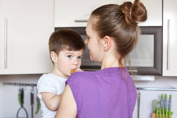 Attractive little male kid eats pastry, look thoughtfully, being on mother`s hands, stand together at kitchen interior, enjoy togetherness. Mum with son at home. Children and eating concept. - Photo, Image