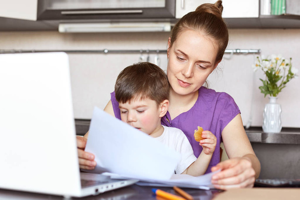 Mother freelancer on maternity leave works with documentation, makes business report on portable laptop computer, holds son who interrupts her, sit together against kitchen interior. Family concept - Foto, afbeelding