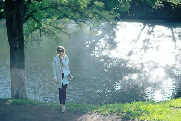 beautiful blonde woman wearing stylish blue coat, walking near lake in park - Photo, image