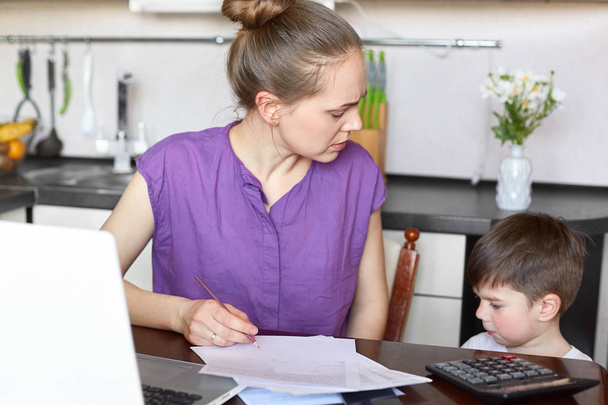 Busy female and young mother makes financial report, works with documents and modern laptop computer, has talk with her small son who feel bored, stands near table in kitchen. Motherhood concept - Photo, Image