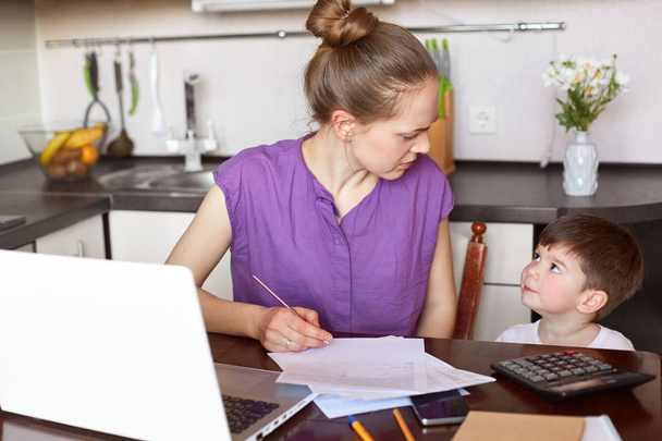 Horizontal shot of working mum being busy with documentation, makes financial report or calculates family budget, surrounded with papers and laptop, looks at her child and asks to be obedient - Φωτογραφία, εικόνα