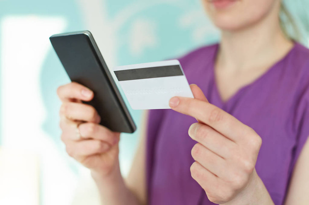 Cropped image of modern smart phone and plastic card in woman`s hands against blue blurred background. Young businesswoman checks her bank account in mobile application. Online payment concept - Foto, Bild
