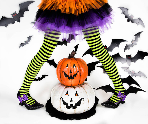 lustige grün schwarz gestreifte Beine eines kleinen Mädchens mit Halloween-Kostüm einer Hexe mit Hexenschuhen und Smiley-Halloween-Kürbis-Buchse o Laterne - Foto, Bild