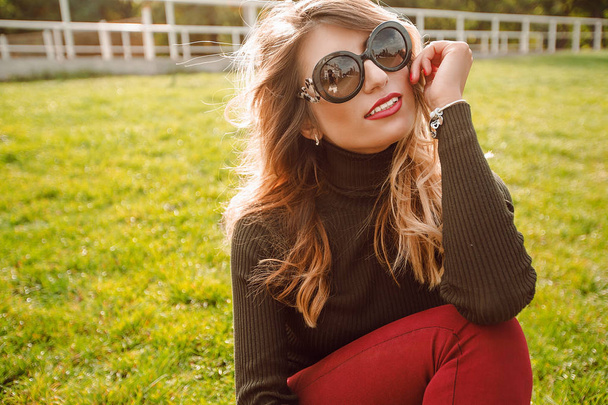 portret uśmiechający się młoda kobieta, noszenie okularów przeciwsłonecznych, siedząc na zielonej trawie w parku  - Zdjęcie, obraz