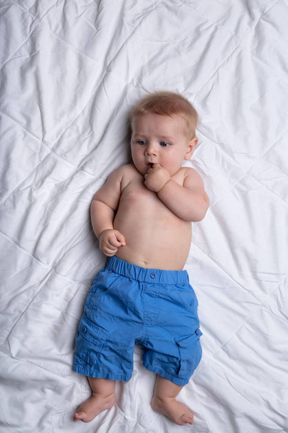 Thoughtful contented little baby boy in over sized blue pants lying on his back sucking a thumb against white bedclothes viewed from above - Photo, Image
