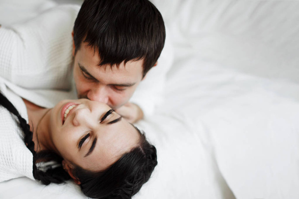 Jovem marido e mulher de casacos brancos brincam e sorriem na cama em um quarto de hotel. Fechar
. - Foto, Imagem