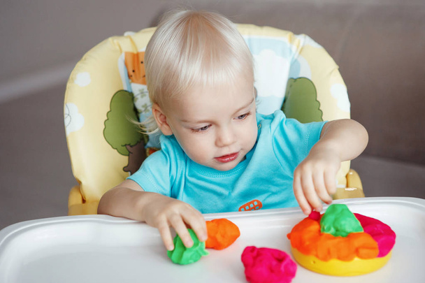 Netter Junge mit blonden Haaren und blauen Augen, der mit Knetmasse spielt und mit rosa, roten und grünen Tassen am heimischen Tisch sitzt. - Foto, Bild