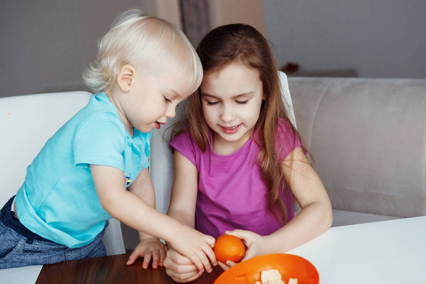 Two happy children Blond boy and girl eats orange, playing sitting at table at home - Zdjęcie, obraz
