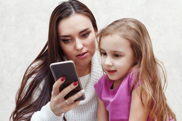 Young woman and girl are looking together at the smartphone. They are smilling and sitting on the sofa. It's mom and daughter. - Photo, image