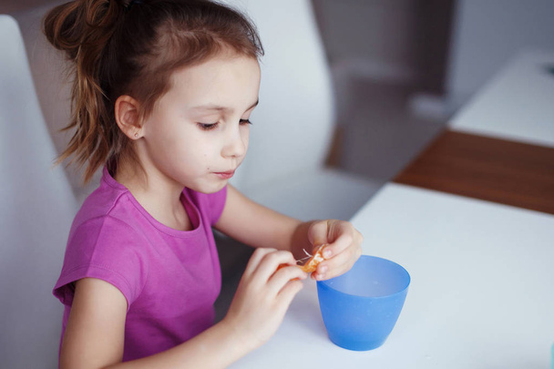 Een meisje zit op een witte tafel. Ze drinkt uit een blauw kopje en eet van cookies. - Foto, afbeelding