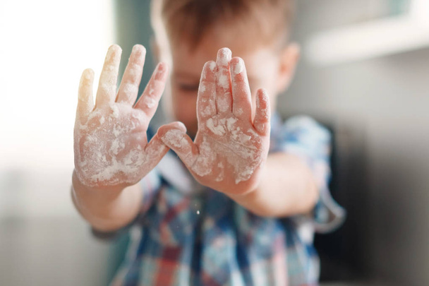 Roztomilý mladý chlapec v kostkované košili vztáhl ruce vpřed s dlaněmi špinavé s moukou - Fotografie, Obrázek