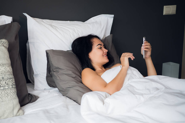 Hermosa mujer joven cubierta por una manta está acostada en la cama con teléfono inteligente
 - Foto, Imagen