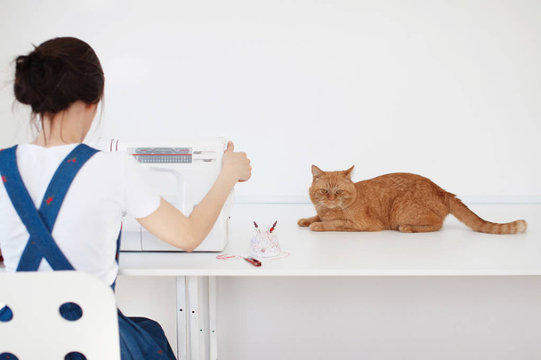 Pretty girl in a blue dress sews on a sewing machine, a red cat sits next to her and watches - 写真・画像