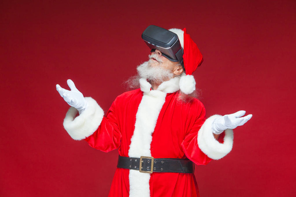 Christmas. Santa Claus in black virtual reality glasses makes gestures with his hands. Surprise, emotion. New technology. Isolated on red background. - Photo, Image