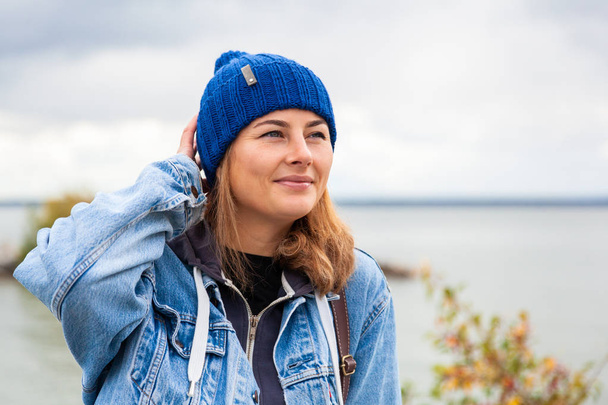 Portrait  outdoor atmospheric lifestyle photo of young beautiful  darkhaired woman  in knitting hat, in a denim jacket and black trousers against the background sea  in sunny autumn day   - Photo, Image