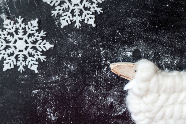 Χριστουγεννιάτικη ιστορία με ένα αρνί. Παιχνίδι αρνί από φυσικό μαλλί και δύο λευκές νιφάδες χιονιού σε ένα σκοτεινό backgroun - Φωτογραφία, εικόνα