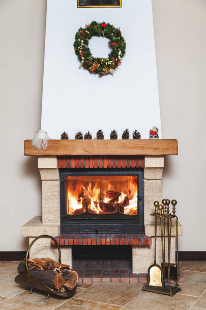 cheminée moderne ornée d'une couronne de Noël et d'une statue du Père Noël. Devant la cheminée, il y a une drovnitsa forgée et un ensemble d'outils en laiton
 - Photo, image