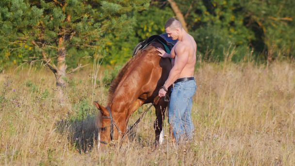 Egy erős férfi meztelen felsőtestét és farmer nadrág áll egy ló, hogy füvet rág - Fotó, kép