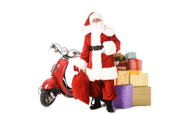 Санта Клаус стоит с винтажным красным скутером, мешок и подарочные коробки и глядя на камеру изолированы на белом
 - Фото, изображение