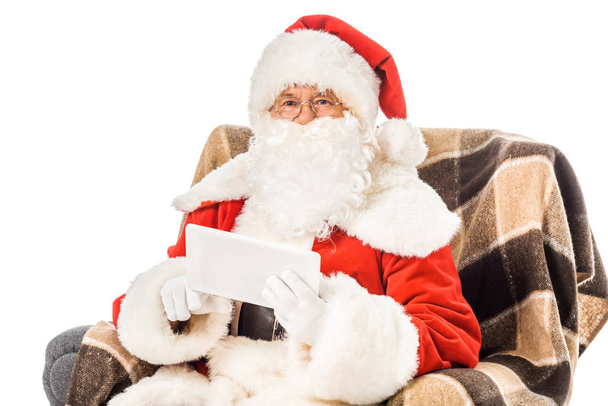 santa claus sitting in armchair with tablet and looking at camera isolated on white - Photo, Image