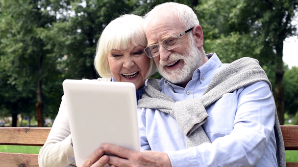 feliz pareja de ancianos usando tableta y saludando las manos durante la videollamada en el parque
 - Metraje, vídeo