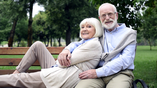 starší pár objímat na lavičce v parku a při pohledu na fotoaparát - Záběry, video